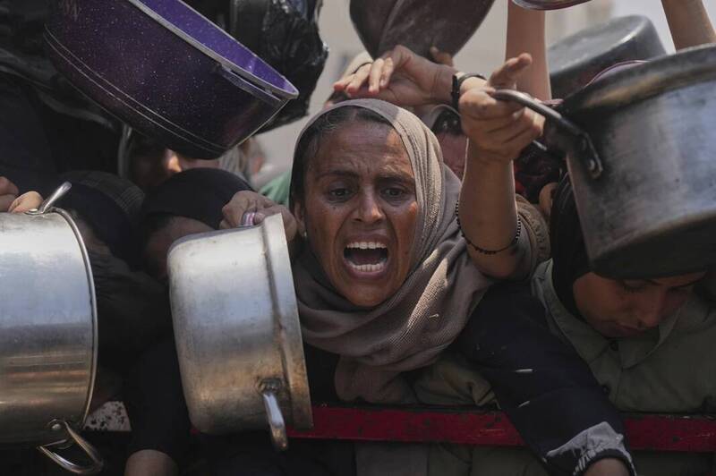 加薩民眾在社區廚房排隊搶領捐贈食物，一名婦女情緒激動，高舉鍋具吶喊求援，反映飢餓壓力與援助混亂場景。（美聯社）
